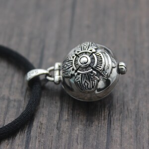 May include: A silver pendant necklace with a round, ornate charm featuring a detailed design. The charm is attached to a black cord.