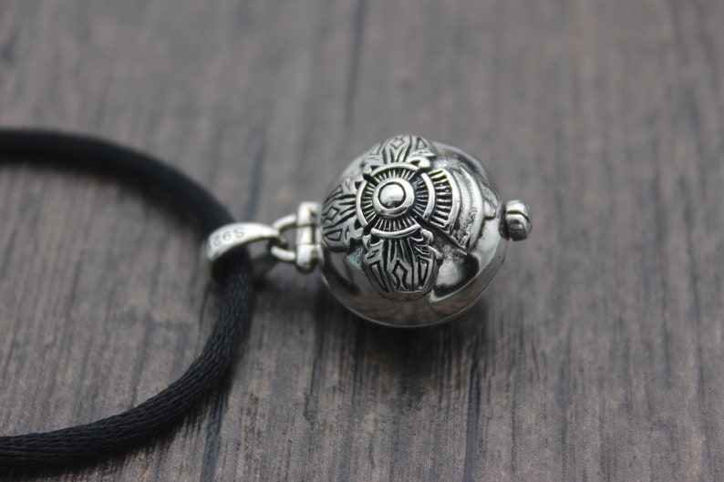May include: A silver pendant necklace with a round, ornate charm featuring a detailed design. The charm is attached to a black cord.
