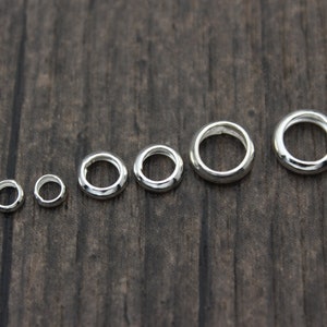 May include: A set of seven silver jump rings of varying sizes, arranged in order from smallest to largest on a wooden surface.
