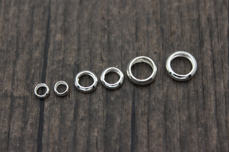 May include: A set of seven silver jump rings of varying sizes, arranged in order from smallest to largest on a wooden surface.