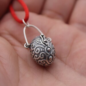 May include: A silver pendant in the shape of a small, ornate, egg-shaped box with a hinged lid. The pendant is attached to a red cord.