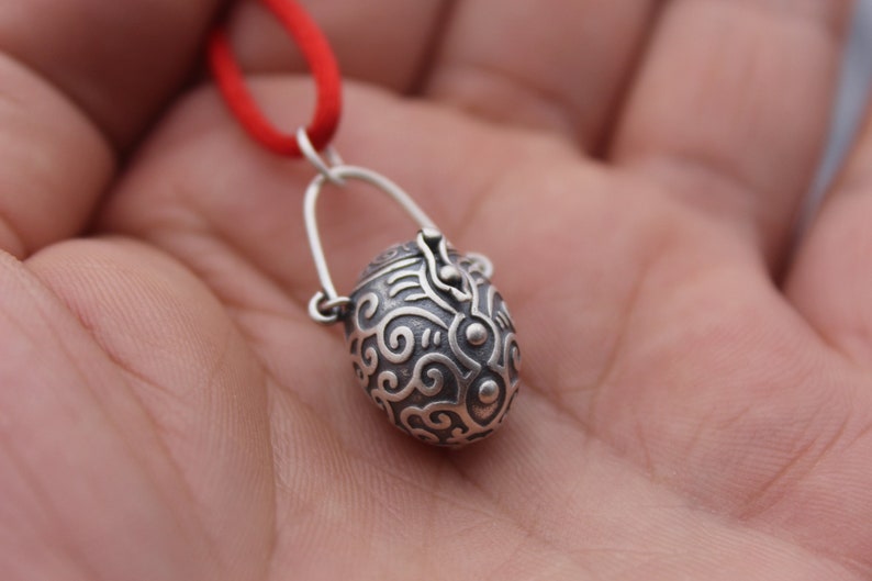 May include: A silver pendant in the shape of a small, ornate, egg-shaped box with a hinged lid. The pendant is attached to a red cord.