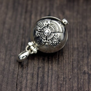 May include: A silver charm with an intricate floral design. The charm is round and has a small loop at the top for attaching to a necklace or bracelet.