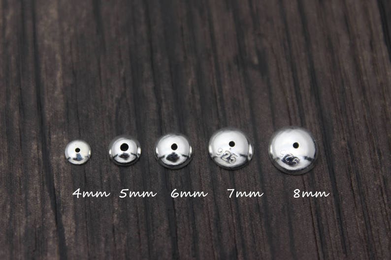 May include: Five silver-toned metal bead caps in different sizes, ranging from 4mm to 8mm. The bead caps are round and have a small hole in the center. The bead caps are arranged in a row from smallest to largest.