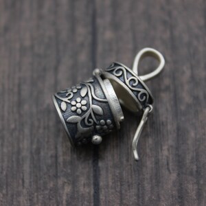 May include: A silver charm with a floral design and a hinged lid. The charm is open and the inside is visible. The charm is on a wooden surface.