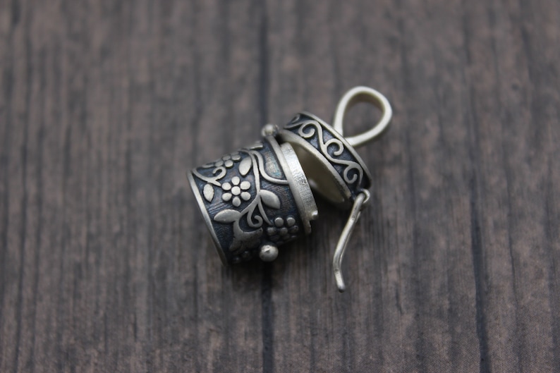 May include: A silver charm with a floral design and a hinged lid. The charm is open and the inside is visible. The charm is on a wooden surface.