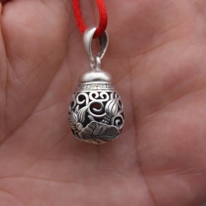 Puede incluir: Un colgante de plata con intrincados dise&ntilde;os florales y una cuerda roja atada. El colgante tiene forma de l&aacute;grima y tiene un centro hueco.