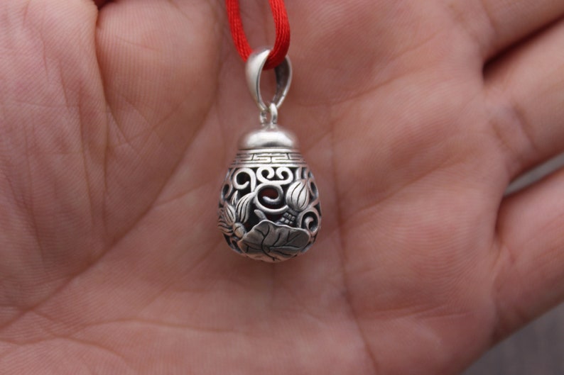 Puede incluir: Un colgante de plata con intrincados dise&ntilde;os florales y una cuerda roja atada. El colgante tiene forma de l&aacute;grima y tiene un centro hueco.