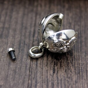 May include: A silver charm in the shape of a sphere with intricate designs. The charm is open and has a small screw on the side. The charm is on a wooden surface.