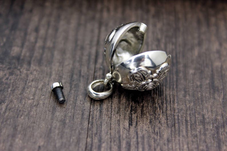 May include: A silver charm in the shape of a sphere with intricate designs. The charm is open and has a small screw on the side. The charm is on a wooden surface.