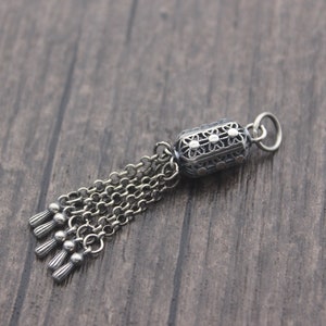 May include: A silver charm with a detailed filigree design and a tassel of chains. The charm is shaped like a cylinder and has a small loop at the top for attaching to a bracelet or necklace.