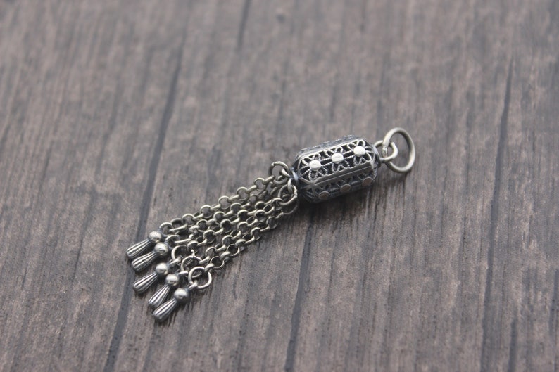 May include: A silver charm with a detailed filigree design and a tassel of chains. The charm is shaped like a cylinder and has a small loop at the top for attaching to a bracelet or necklace.