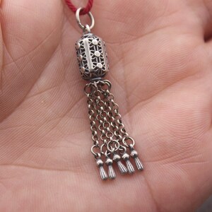 May include: A silver charm with a filigree design and a tassel. The charm is attached to a red string.