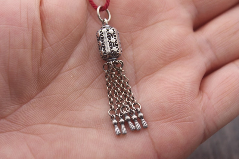 May include: A silver charm with a filigree design and a tassel. The charm is attached to a red string.