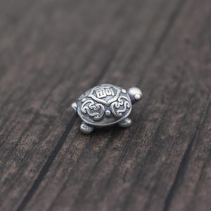 May include: A small silver turtle charm featuring intricate details and Chinese characters on its shell.