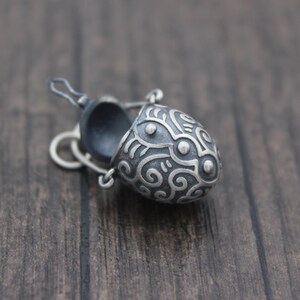 May include: A small, silver, egg-shaped pendant with an intricate, swirling design. The pendant is open and has a black interior.