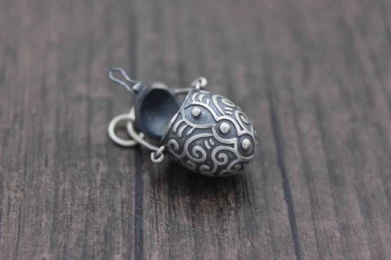 May include: A small, silver, egg-shaped pendant with an intricate, swirling design. The pendant is open and has a black interior.