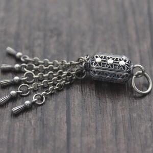 May include: A silver charm with a detailed filigree design and a dangling chain with five tassels. The charm has a small loop for attaching to a bracelet or necklace.