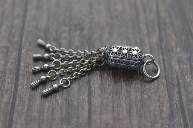 May include: A silver charm with a detailed filigree design and a dangling chain with five tassels. The charm has a small loop for attaching to a bracelet or necklace.