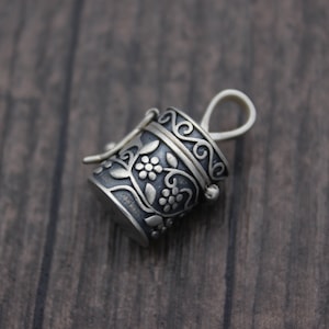 May include: A silver charm in the shape of a small, ornate box with a floral design. The box has a hinged lid and a loop for attaching to a necklace or bracelet.