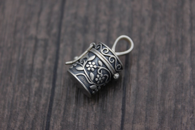 May include: A silver charm in the shape of a small, ornate box with a floral design. The box has a hinged lid and a loop for attaching to a necklace or bracelet.