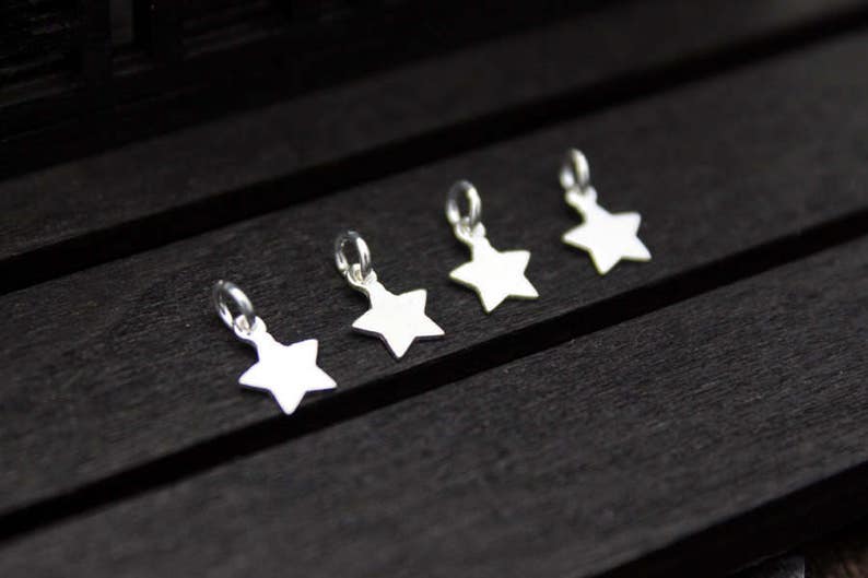 May include: Five silver star-shaped charms with jump rings. The charms are arranged in a row on a black wooden surface.