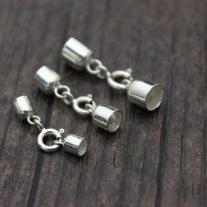 May include: Three silver-colored jewelry clasps with a cylindrical design and a small chain. The clasps are arranged on a dark wooden surface. The clasps have a shiny, metallic finish and are designed for jewelry making.