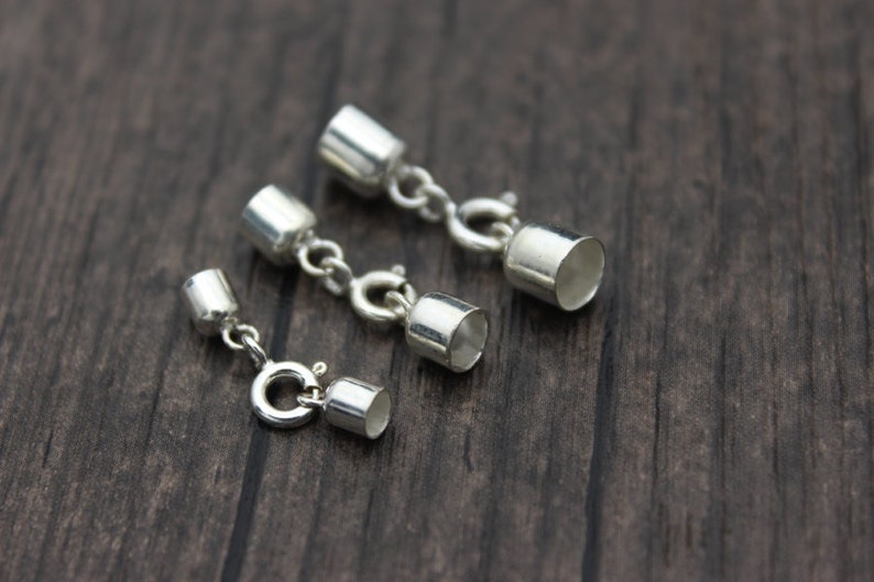 May include: Three silver-colored jewelry clasps with a cylindrical design and a small chain. The clasps are arranged on a dark wooden surface. The clasps have a shiny, metallic finish and are designed for jewelry making.