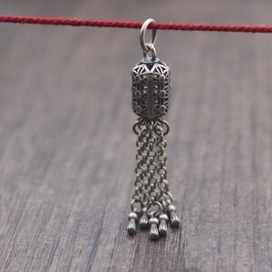 May include: A silver pendant with a detailed, ornate design and a tassel of chains. The pendant is hanging from a red string.