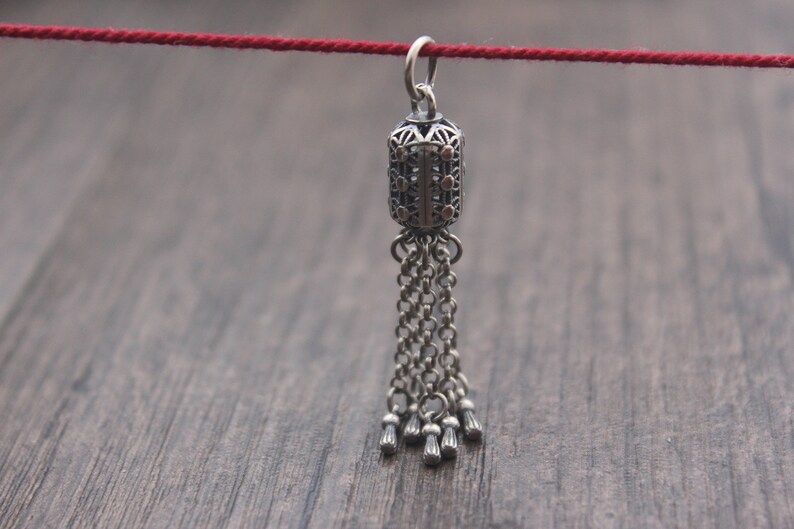 May include: A silver pendant with a detailed, ornate design and a tassel of chains. The pendant is hanging from a red string.