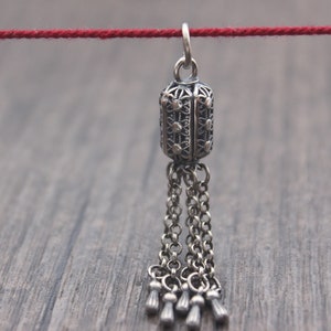 May include: A silver pendant with a filigree design and a tassel of chains. The pendant is hanging from a red string.