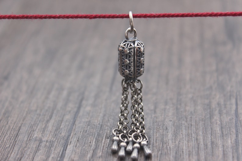 May include: A silver pendant with a filigree design and a tassel of chains. The pendant is hanging from a red string.