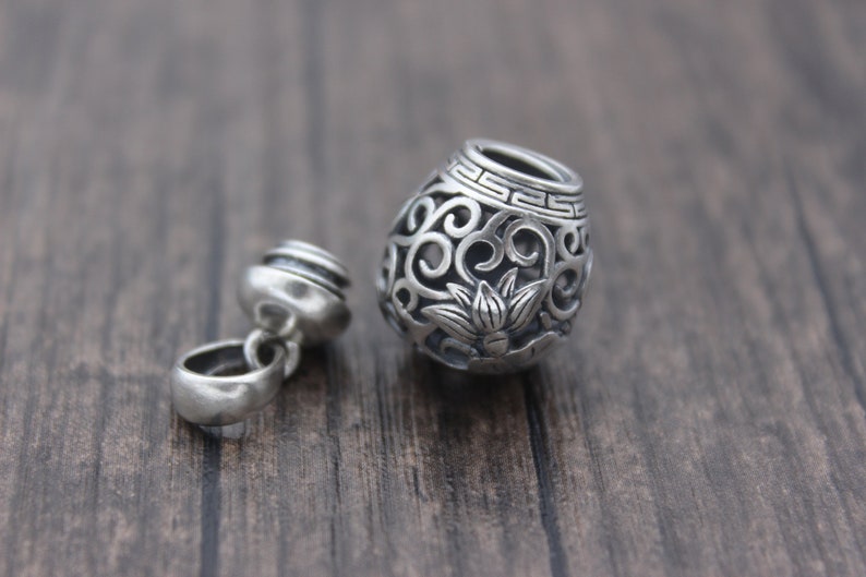 Puede incluir: Un charm de plata con forma de jarr&oacute;n peque&ntilde;o y ornamentado con un dise&ntilde;o de flor de loto. El charm est&aacute; abierto y tiene un peque&ntilde;o bucle en el lateral para sujetarlo a un collar o pulsera.