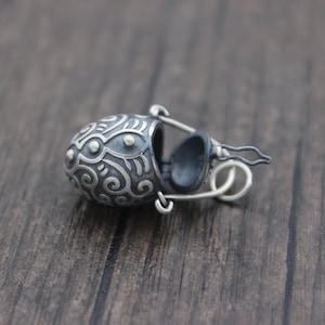 May include: A silver pendant in the shape of a small, ornate, hinged box with a detailed design. The box is open and the inside is black.