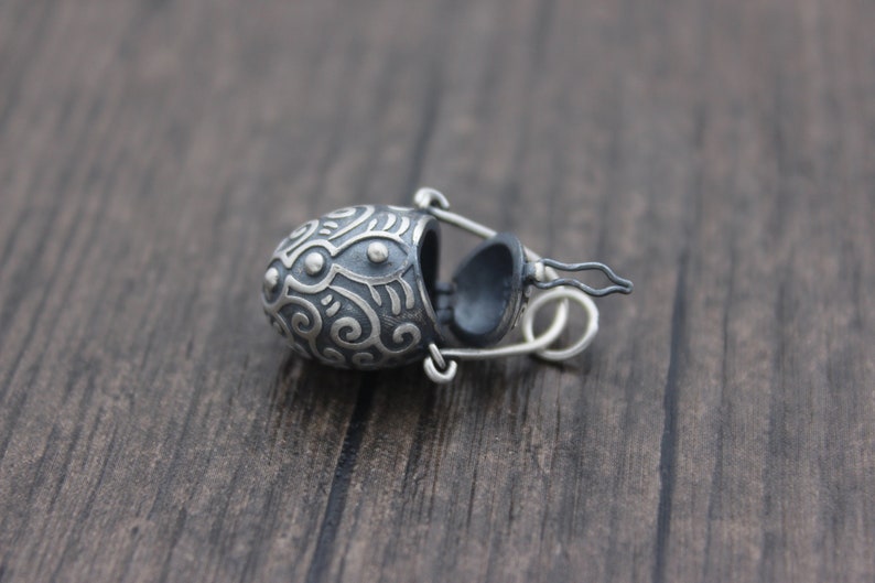 May include: A silver pendant in the shape of a small, ornate, hinged box with a detailed design. The box is open and the inside is black.