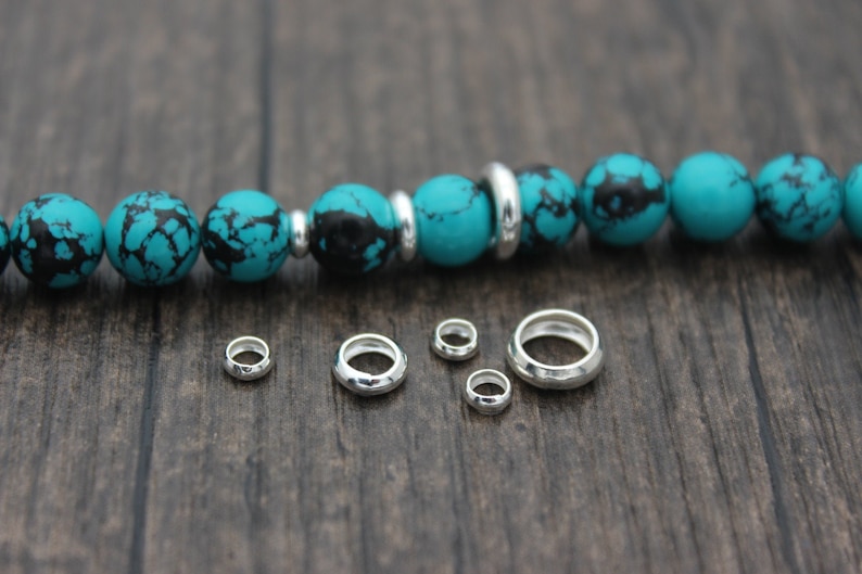 May include: Five silver jump rings of varying sizes on a wooden surface. The jump rings are used for jewelry making.