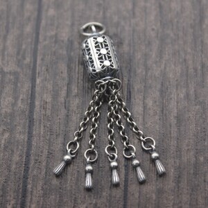 May include: A silver charm with a decorative filigree design and five dangling tassels. The charm is a cylinder shape with a small loop at the top for attaching to a bracelet or necklace.