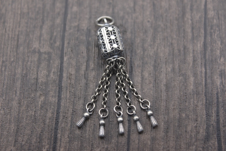 May include: A silver charm with a decorative filigree design and five dangling tassels. The charm is a cylinder shape with a small loop at the top for attaching to a bracelet or necklace.