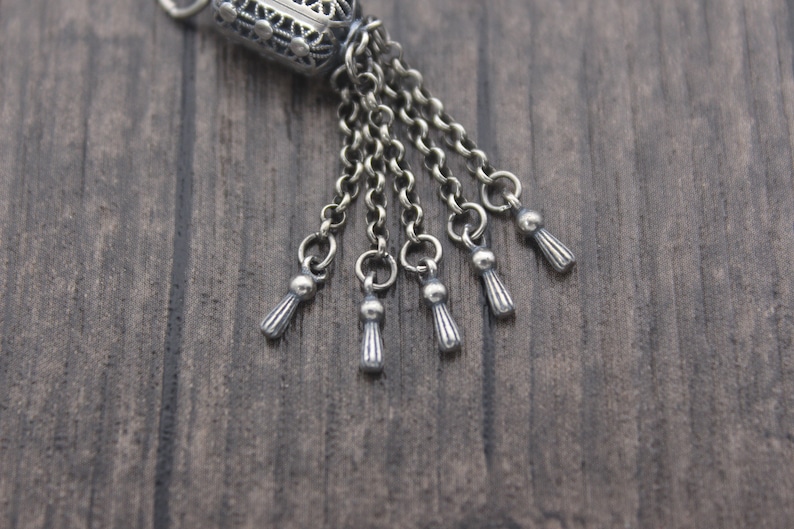 May include: Silver charm with a detailed filigree design and multiple dangling chains with small tassel-like ends.