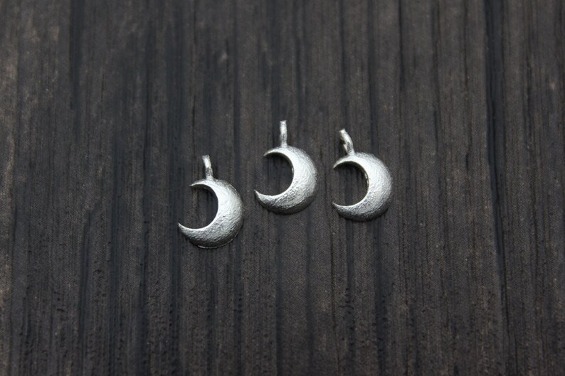 May include: Three silver crescent moon charms on a dark wood background.