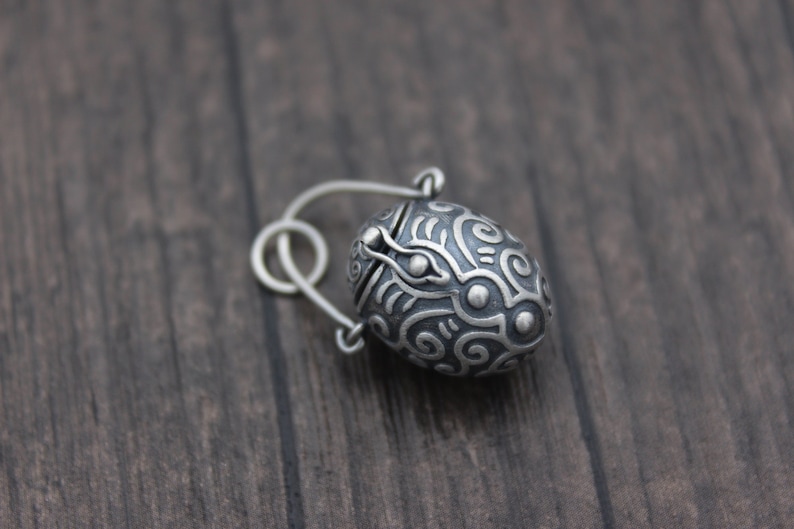 May include: A silver charm in the shape of an egg with an intricate design. The egg has a hinged lid and a small loop for attaching to a chain or bracelet.