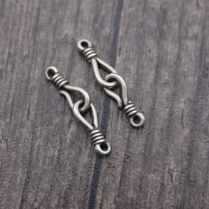 Op de afbeelding: Twee zilverkleurige metalen sieradenconnectoren. Elke connector heeft een lusvormig ontwerp met gewikkelde draaddetails. De connectoren worden getoond op een donker houten oppervlak, ideaal voor het maken van sieraden of knutselen.