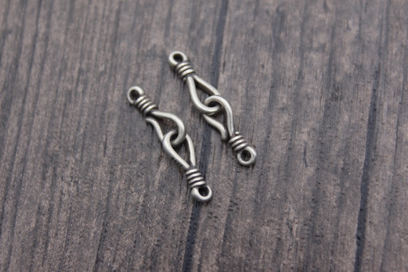 Op de afbeelding: Twee zilverkleurige metalen sieradenconnectoren. Elke connector heeft een lusvormig ontwerp met gewikkelde draaddetails. De connectoren worden getoond op een donker houten oppervlak, ideaal voor het maken van sieraden of knutselen.