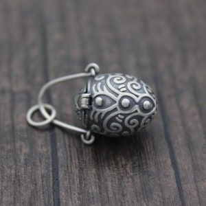 May include: A silver oval-shaped locket with a hinged lid. The locket is decorated with a swirling, intricate pattern and has a small, round, silver charm attached to the top.