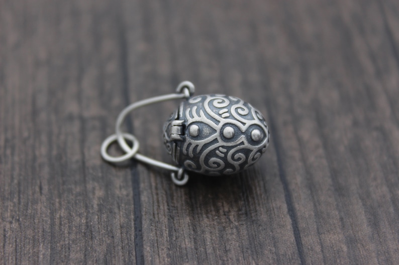 May include: A silver oval-shaped locket with a hinged lid. The locket is decorated with a swirling, intricate pattern and has a small, round, silver charm attached to the top.