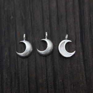 May include: Three silver crescent moon charms with a textured surface. The charms are arranged in a row on a dark wooden background.