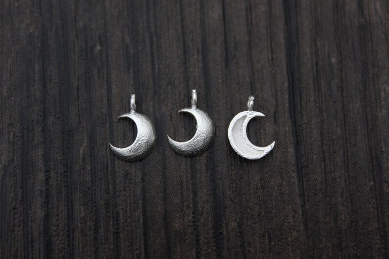 May include: Three silver crescent moon charms with a textured surface. The charms are arranged in a row on a dark wooden background.