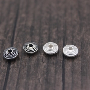 Peut inclure: Quatre perles rondes argentées avec un motif texturé et pointillé. Les perles sont espacées uniformément et ont un trou au centre. Les perles sont sur une surface en bois brun foncé.