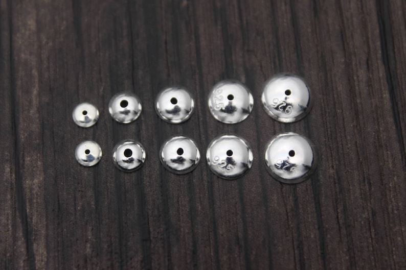 May include: A set of ten silver-toned metal bead caps of varying sizes. The caps are round and have a small hole in the center. The caps are marked with the number "925" indicating they are made of sterling silver.