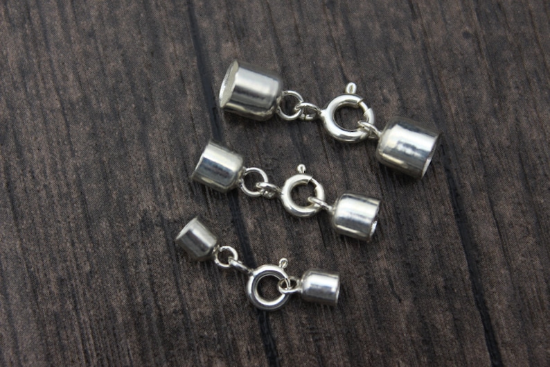 May include: Three silver-colored jewelry clasps with a cylindrical design and a spring ring clasp. The clasps are connected by small chains and are displayed on a dark wood surface.